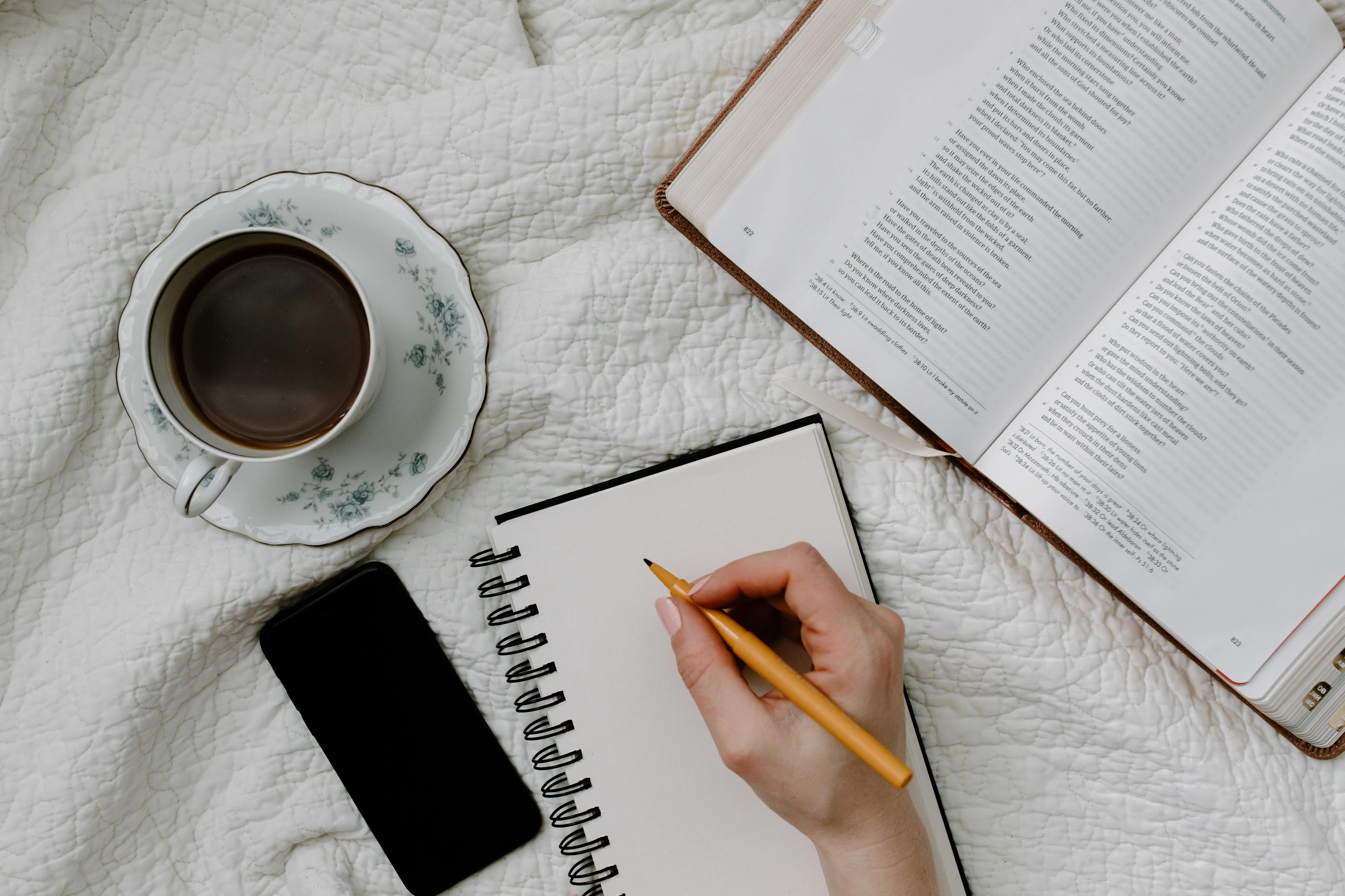 A candid photograph of a person writing in a small notebook beside an open Bible at a simple kitchen table, natural daylight