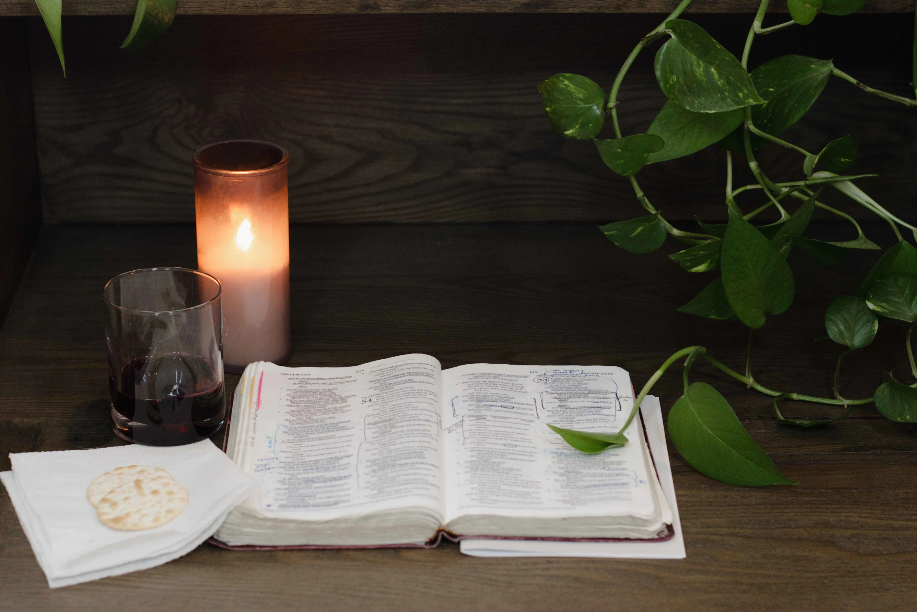 A close photograph of an open Bible on a wooden table beside a lit candle and a mug of tea, in warm evening light