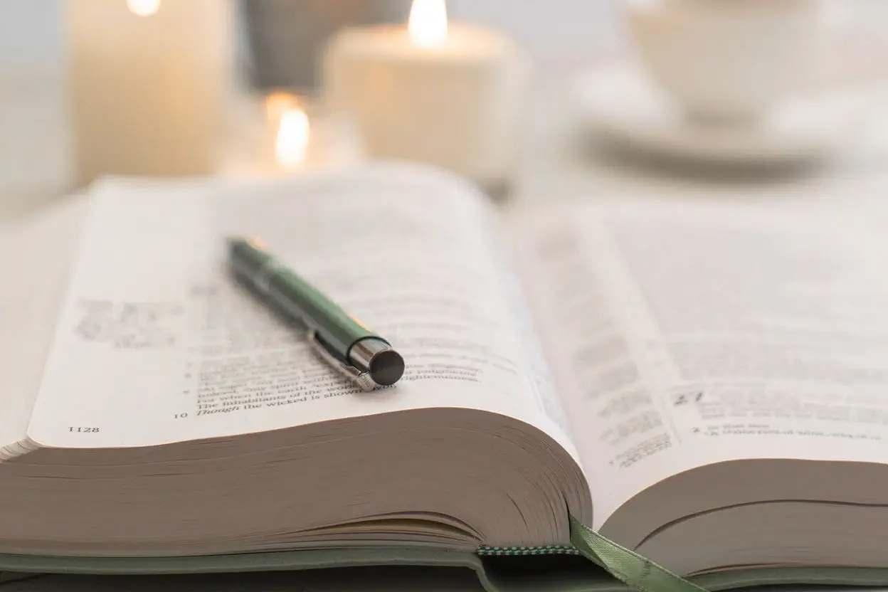 A close-up photograph of an open Bible on a wooden table beside a lit candle and a simple notebook, warm evening light