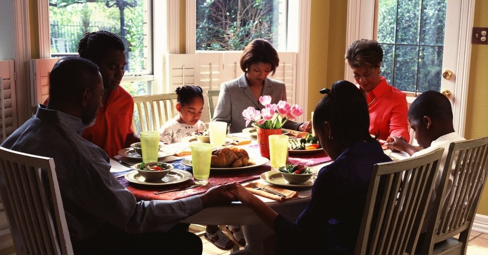 A family seated around a dinner table with heads gently bowed and hands joined, pausing for a brief prayer before eating
