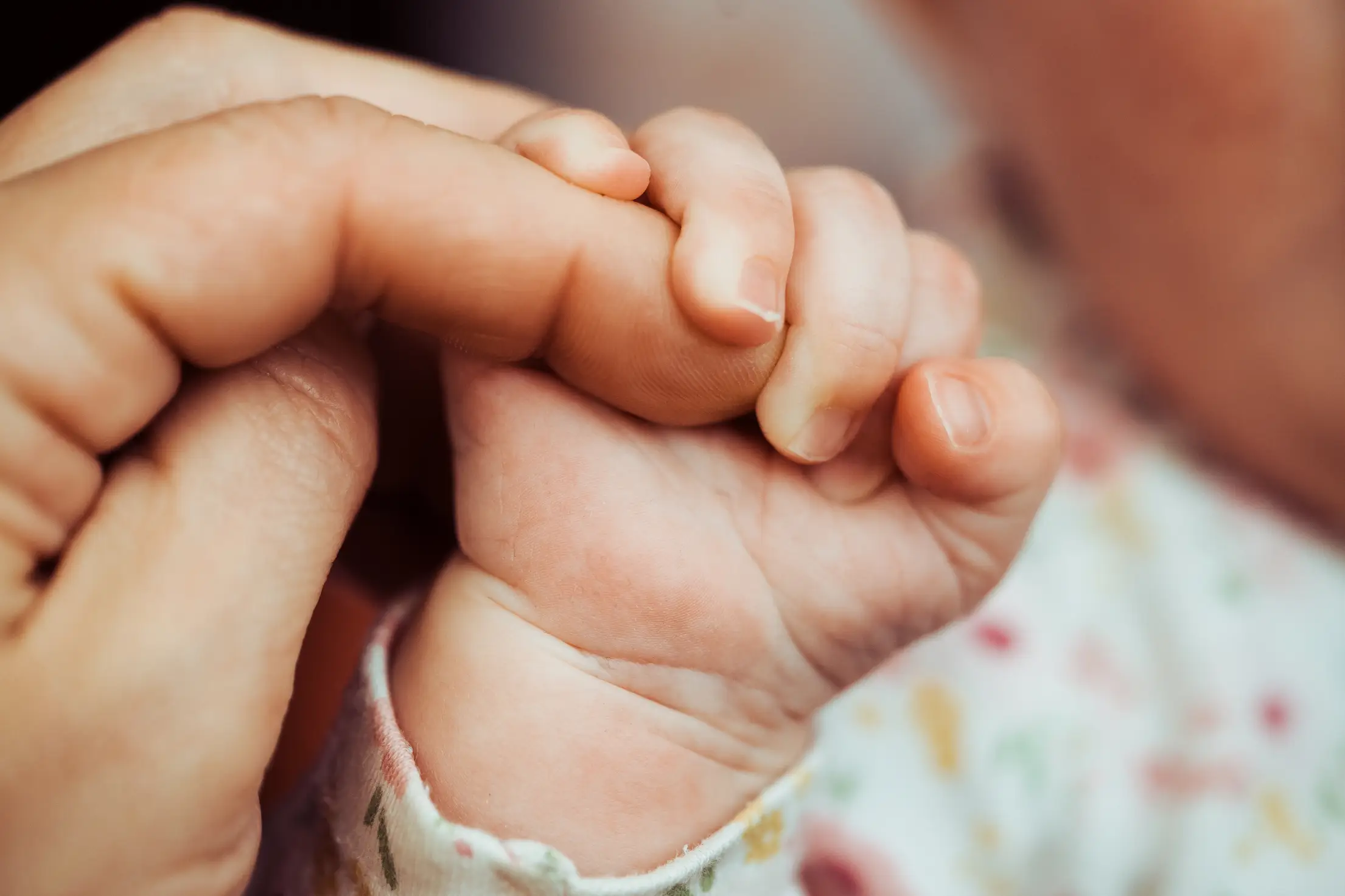 A newborn baby gently holding an adult finger while resting in a parent’s arms in soft window light, intimate home photograph