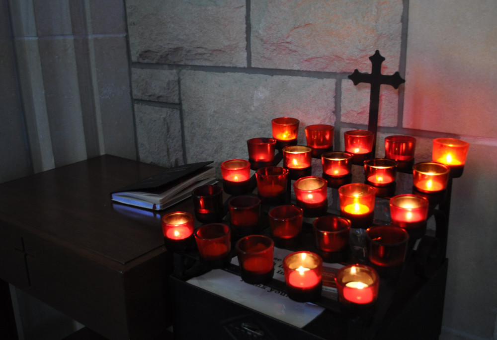 A person lighting a small candle in a dim cathedral, warm candlelight reflecting on stone walls while other votive candles glow softly nearby