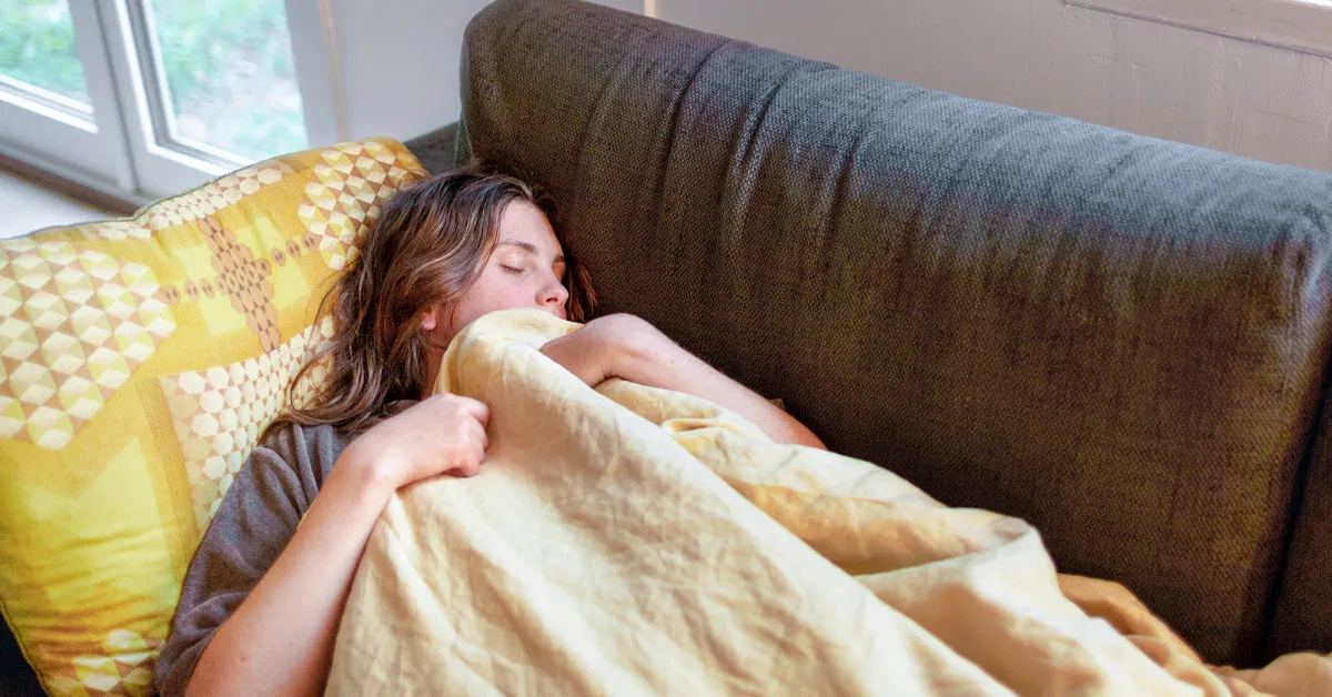 A person resting on a couch near a window in soft morning light, eyes closed, with a blanket pulled up and a calm, unhurried atmosphere