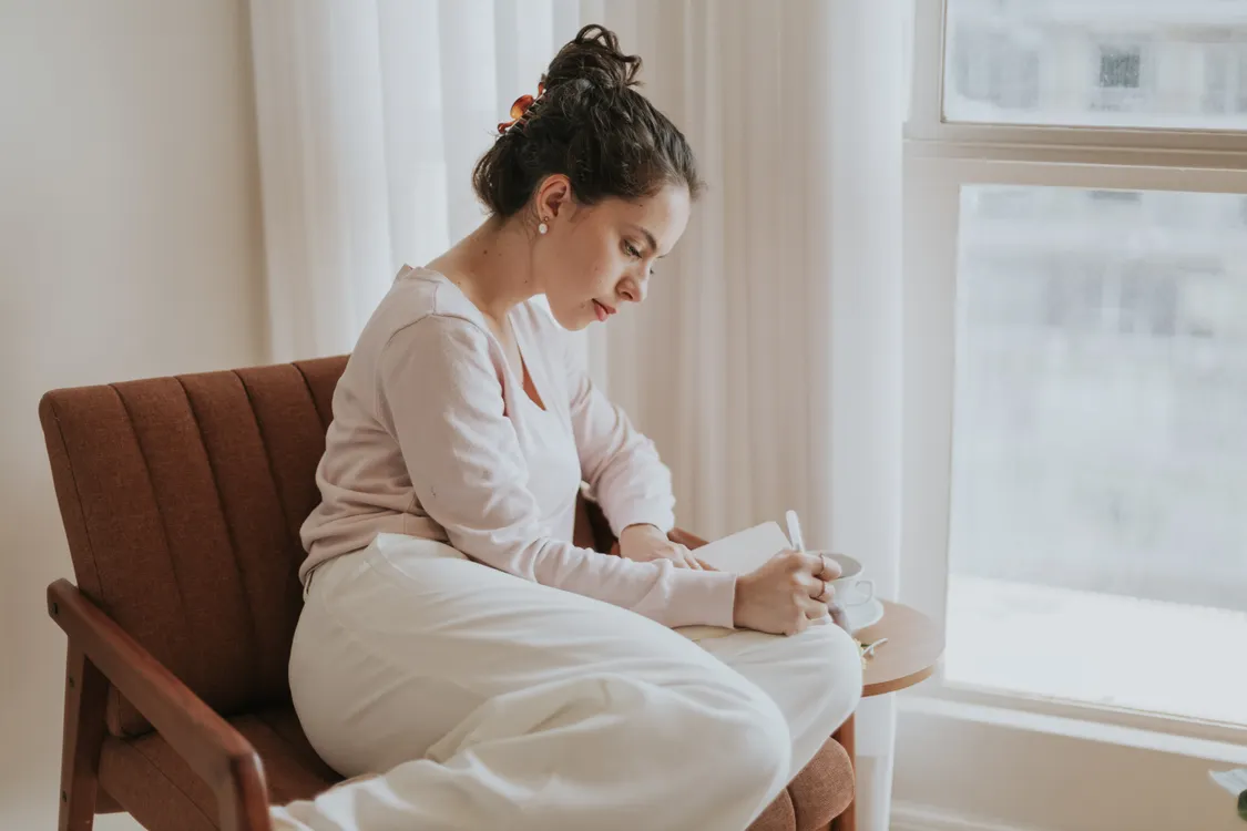 A person seated at a wooden table by a rainy window, writing in a journal beside a mug of tea, quiet morning light