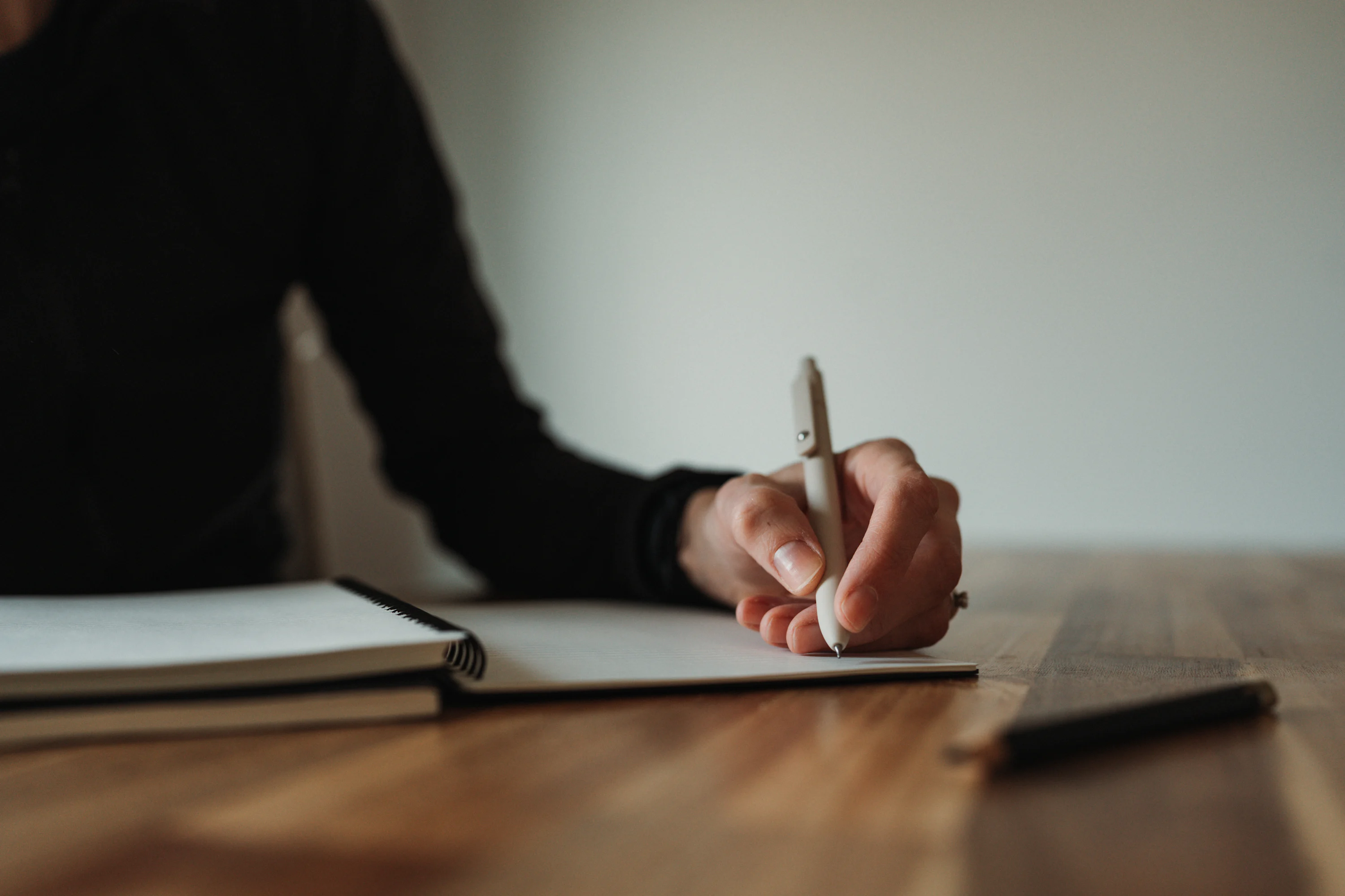 A person sits at a wooden table in the evening, writing in a journal beside a candle and a mug of tea.