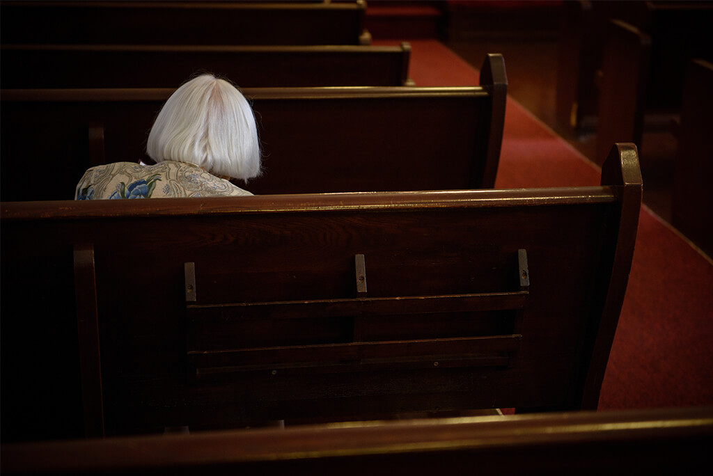 A person sitting alone on a wooden church pew with their hands folded loosely, looking tired and distant as light filters through stained glass