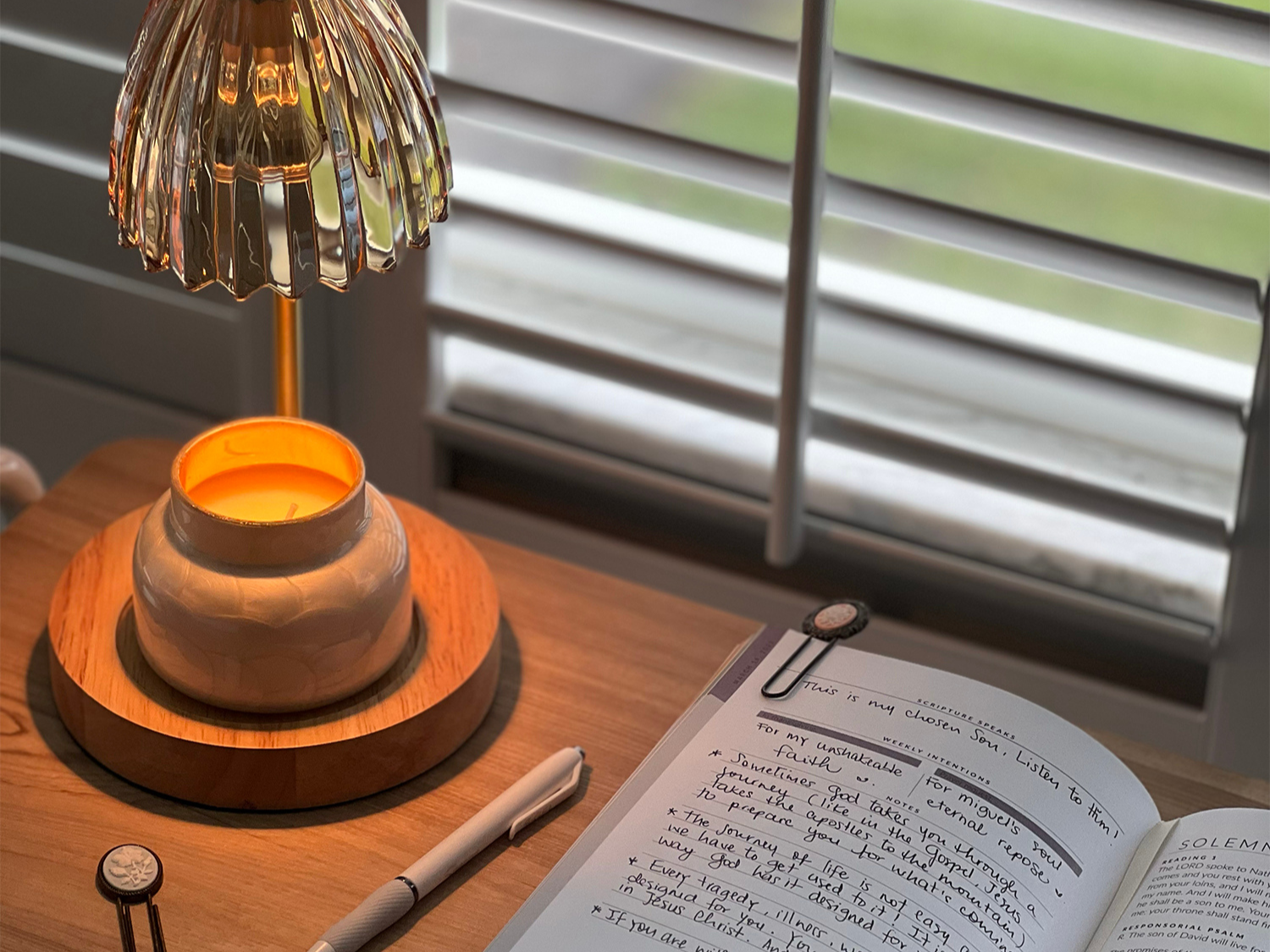 A person sitting at a small table by a window in soft morning light, hands resting near a lit candle and a simple journal, creating a calm space for prayer