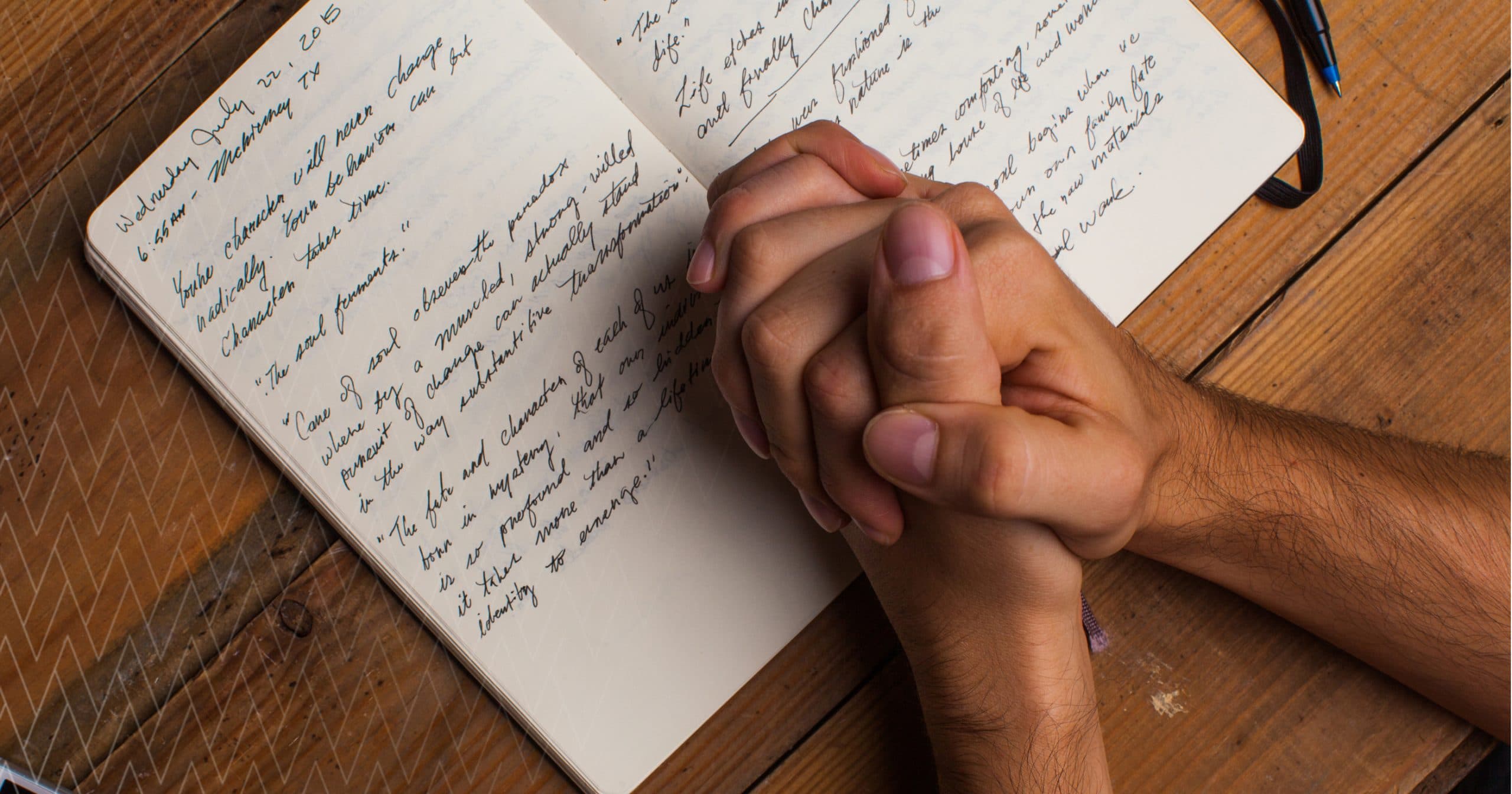 A person sitting by a window writing in a small notebook with a mug beside them, pausing to reflect before prayer