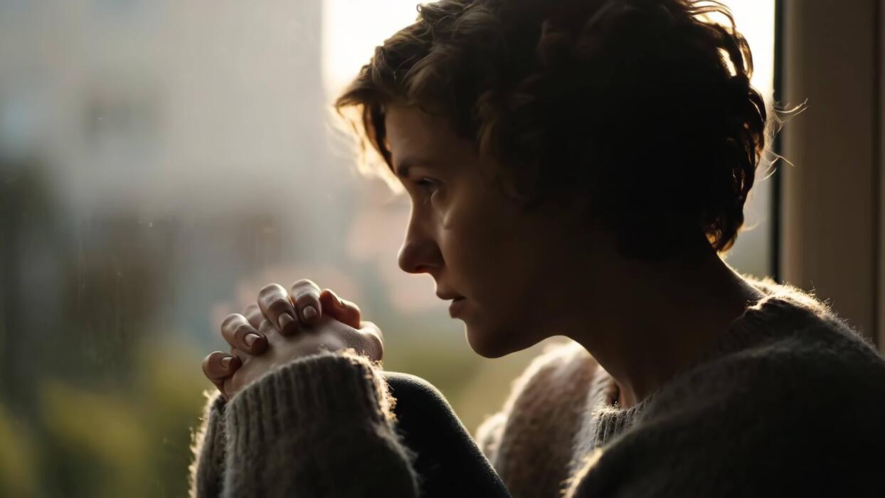 A person sitting on the edge of a bed near a bright window at sunrise, hands gently clasped in quiet prayer