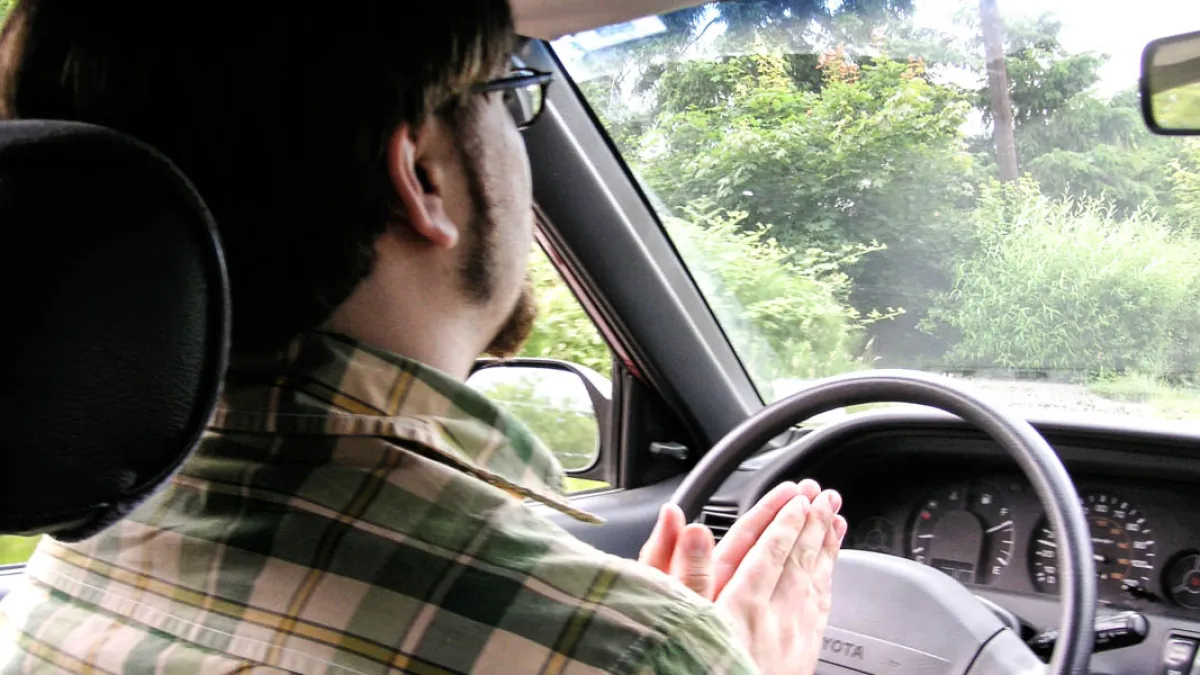 A person sitting quietly in a parked car with hands resting on the steering wheel and eyes closed in a brief moment of prayer during daylight