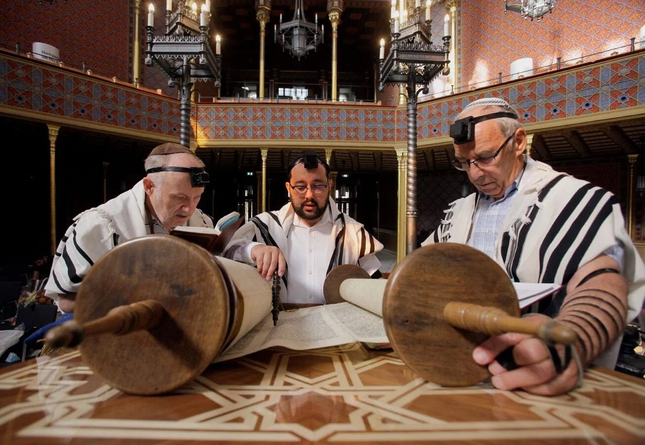 A person standing at a reading table unrolling a large parchment scroll inside an old stone synagogue, warm ambient light.