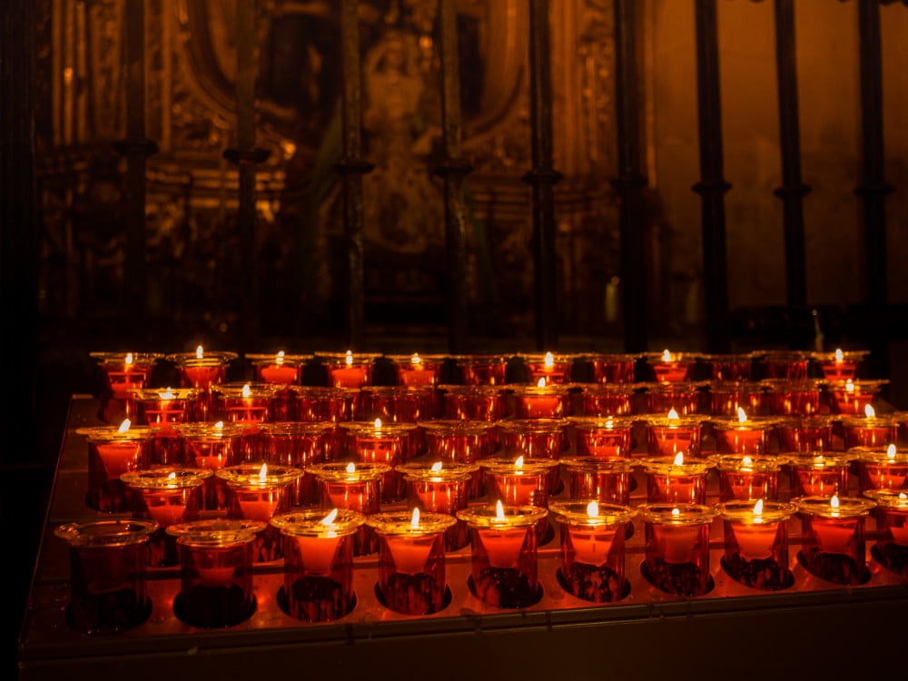 A quiet side chapel in a Catholic church with a row of votive candles glowing and a few empty chairs nearby