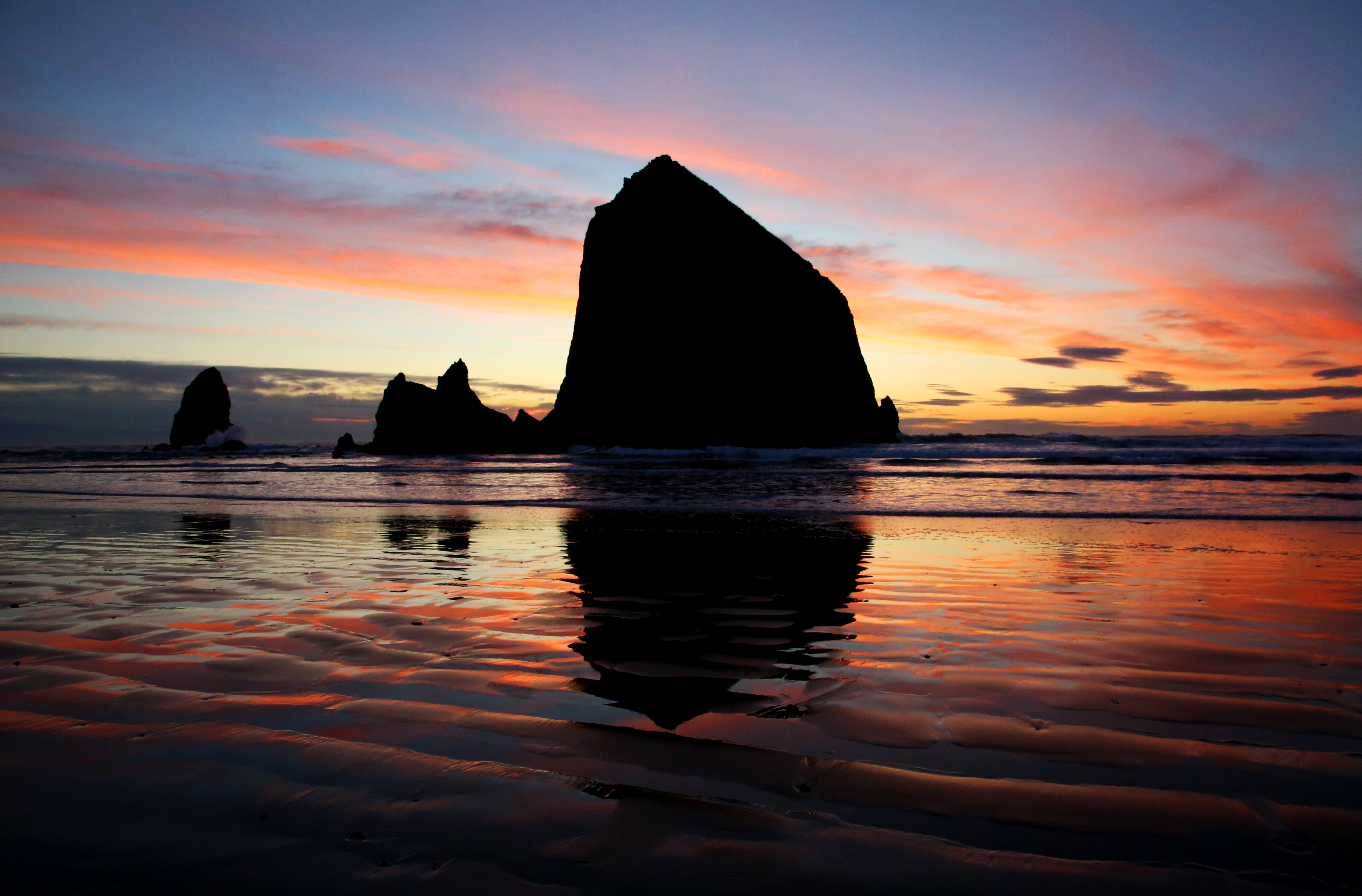 A solitary person sitting on a driftwood log facing the ocean at sunset on the Oregon coast, waves rolling in under a glowing sky