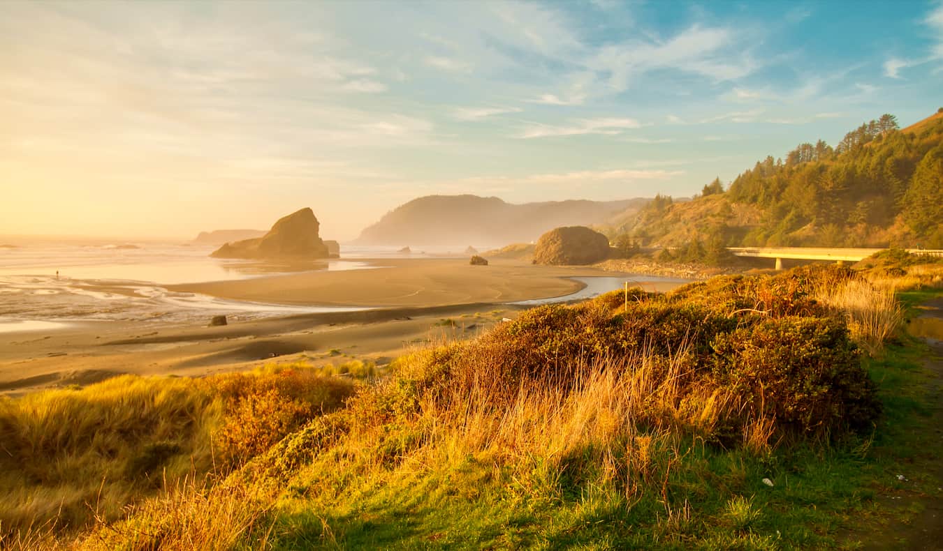 A solitary person sitting quietly on a rocky Oregon beach at sunrise, wrapped in a warm blanket, facing the ocean in a calm moment of reflection