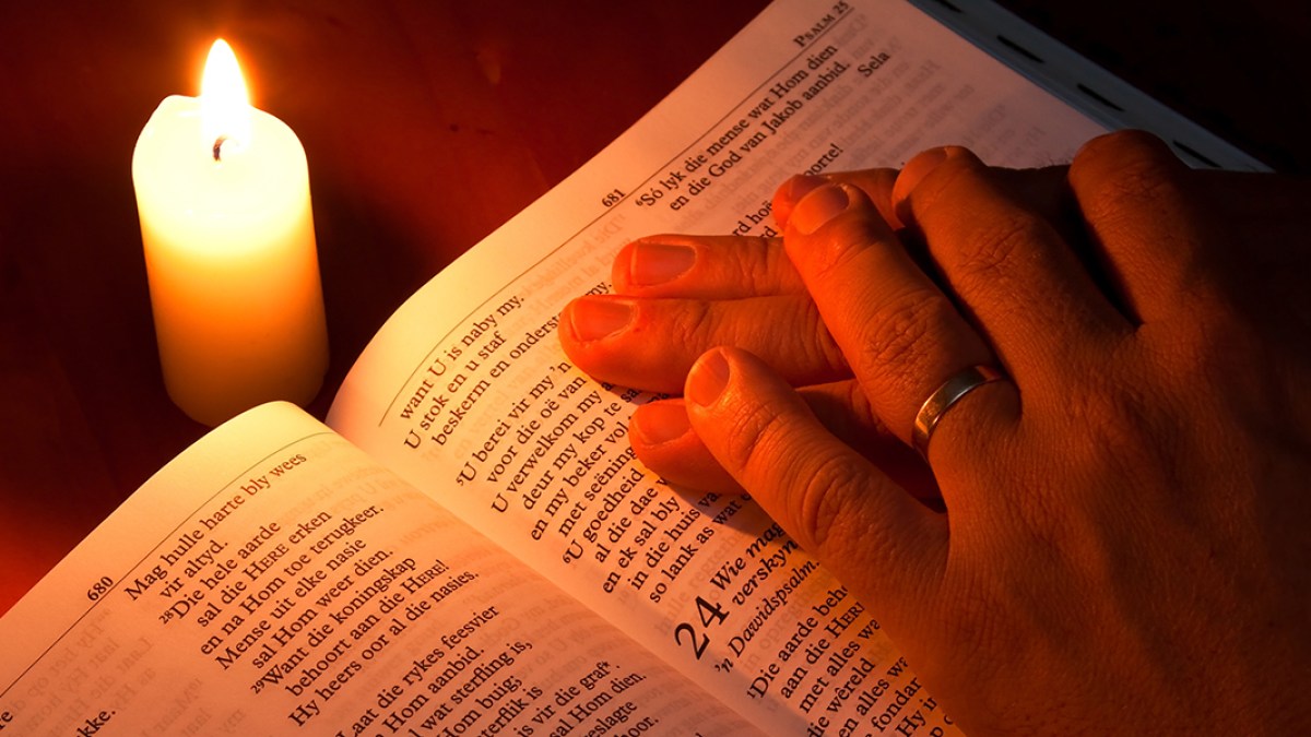 Hands lighting a single candle on a small table at home beside an open book, warm evening light creating a quiet devotional atmosphere