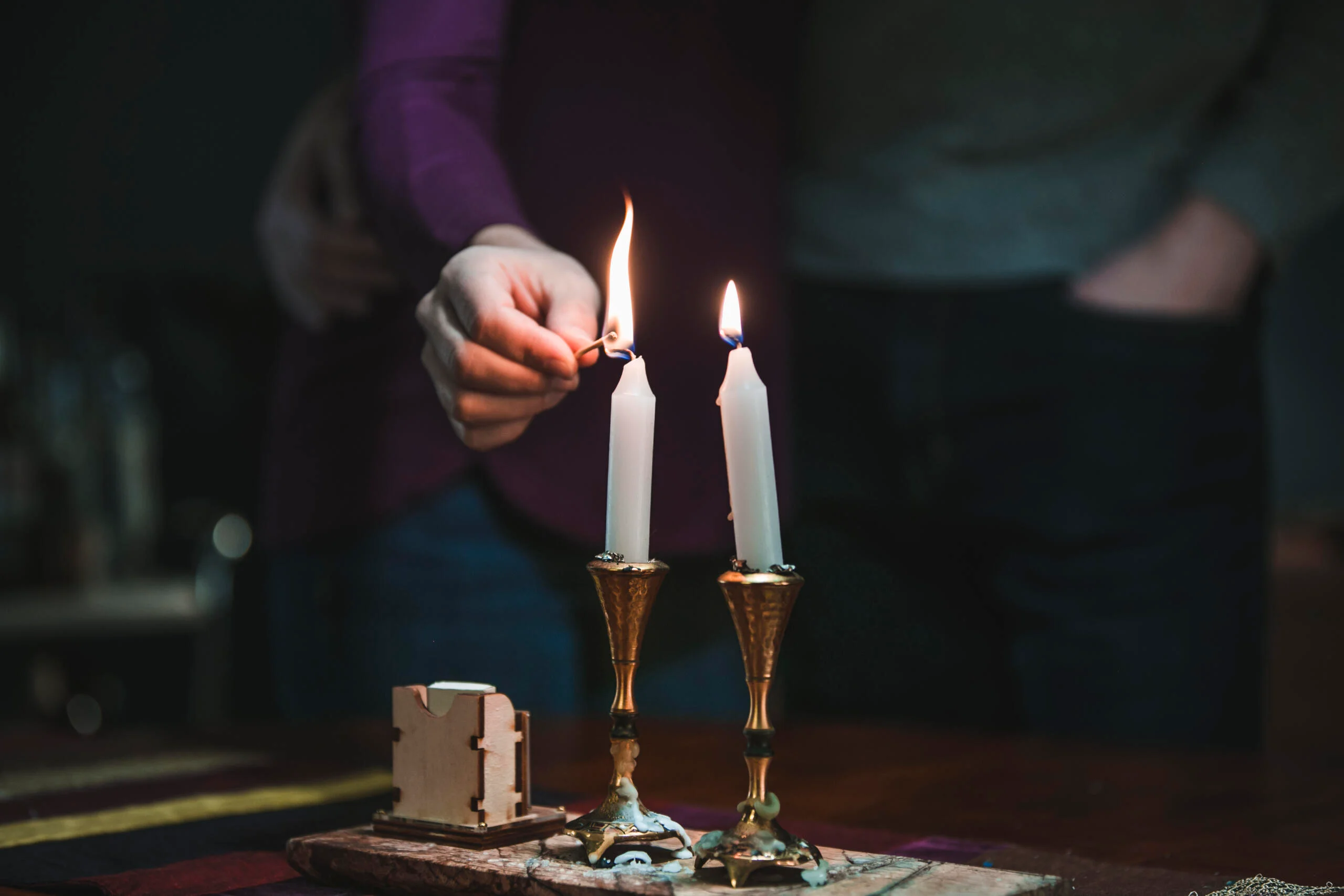 Two lit Shabbat candles on a dining table beside a simple loaf of challah and a folded prayer booklet, photographed in warm evening light