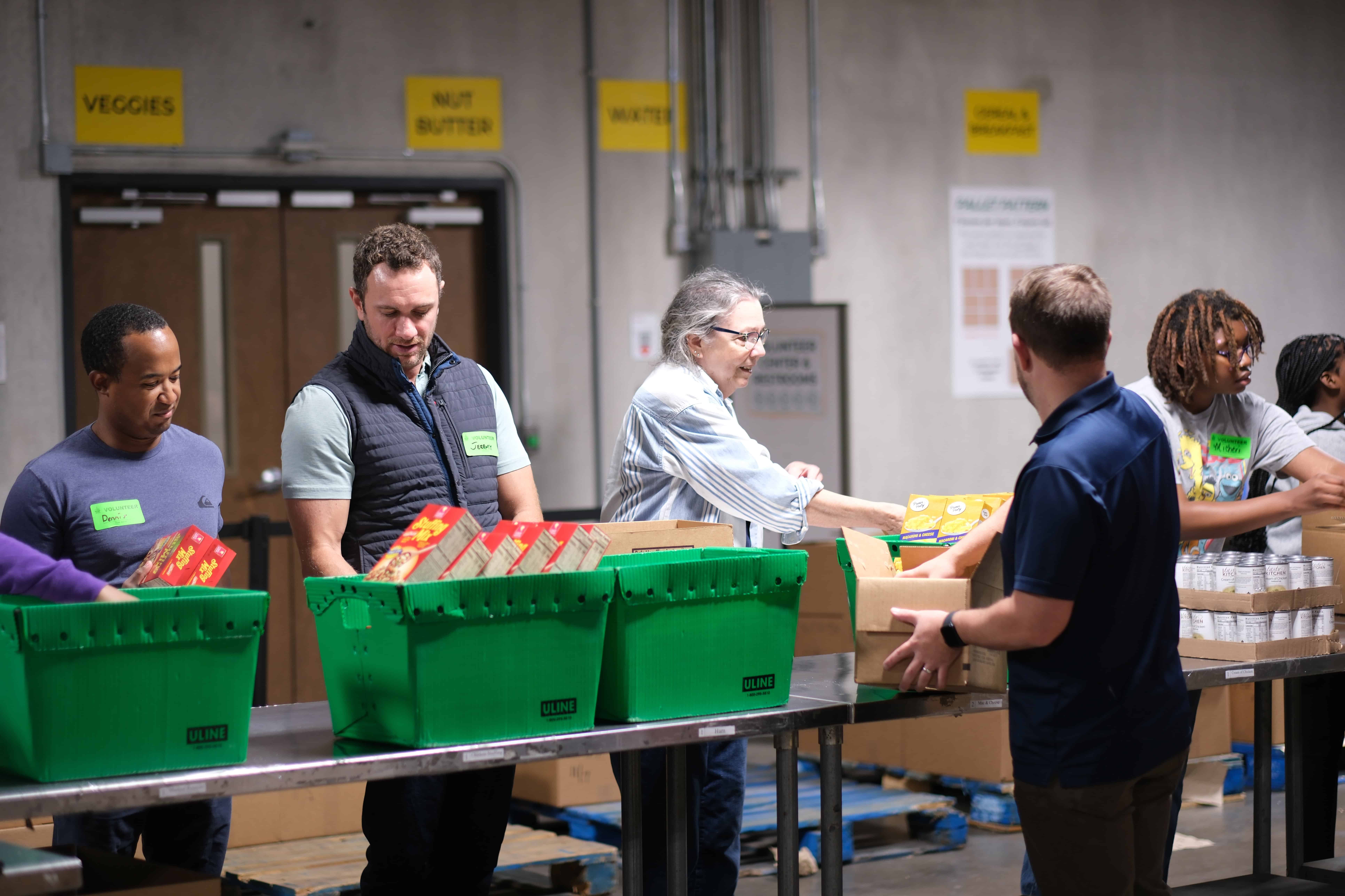 Two volunteers standing at a long table in a food pantry, sorting donated canned goods into boxes, focused and calm in a bright room
